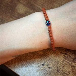 Puede incluir: Un brazalete de cuentas de madera marrón con una cuenta de vidrio azul con puntos multicolores.