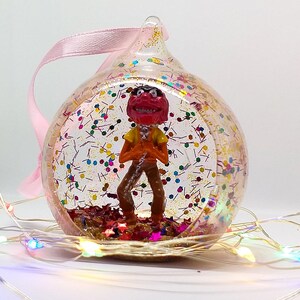 Puede incluir: Un adorno de vidrio transparente con una cinta rosa y una pequeña figura de plástico de un personaje pelirrojo con un micrófono en su interior. El adorno está lleno de purpurina y confeti.