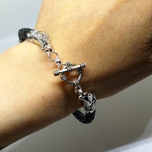 May include: A black braided leather bracelet with silver-toned eagle head accents and a toggle clasp. The bracelet is shown on a wrist, showcasing the intricate details of the jewelry.