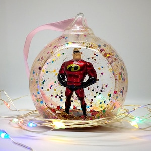 Puede incluir: Un adorno navideño de vidrio transparente con una cinta rosa y una figura de superhéroe roja y negra en su interior. El adorno está lleno de confeti y purpurina de colores.
