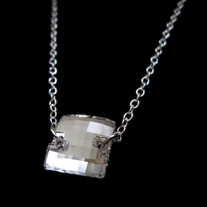 May include: A silver necklace with a square, faceted crystal pendant. The crystal is clear and reflects light, and the chain is delicate. The necklace is displayed against a black background.