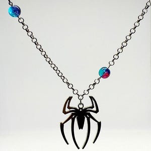 Può includere: Collana color argento con un ciondolo a forma di ragno stilizzato. La catena ha due perline rotonde colorate. Il ciondolo a ragno è nero e presenta un design dettagliato. La collana è esposta su sfondo bianco.
