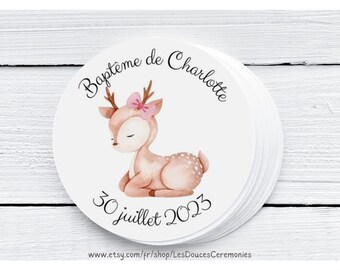 May include: Round sticker with a watercolor illustration of a fawn with a pink bow. The text "Bapteme de Charlotte" and "30 juillet 2023" is printed on the sticker. The sticker is on a white wooden surface.