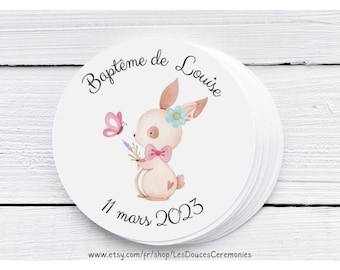 May include: Round sticker with a watercolor illustration of a pink bunny holding a purple flower and a pink butterfly. The text "Bapteme de Louise" and "11 mars 2023" are printed on the sticker. The sticker is on a white wooden surface.