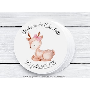 Op de afbeelding: Ronde sticker met een aquarel-illustratie van een fawn met een roze strik. De tekst "Bapteme de Charlotte" en "30 juillet 2023" staat op de sticker gedrukt. De sticker ligt op een wit houten oppervlak.