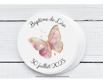 May include: A stack of white circular stickers. Each sticker features a pink and gold butterfly design with the text "Baptême de Lisa" and "30 juillet 2023". The stickers are on a white wooden surface.