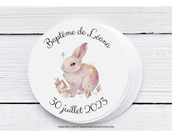 May include: A stack of white round stickers. Each sticker features a watercolor illustration of a pink rabbit with gold accents and the text "Baptême de Léana" and "30 juillet 2023".