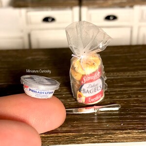 May include: A miniature plastic container of Philadelphia cream cheese next to a bag of mini everything bagels. The bag is tied with a string and has a red and white label that says "Strafel's Everything Bagels".