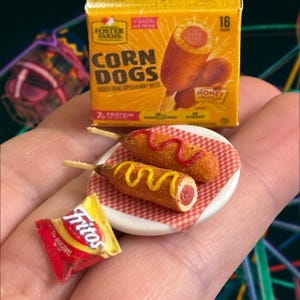 Puede incluir: Un plato blanco en miniatura con un patrón de cuadros rojos y blancos contiene dos mini-perritos calientes de maíz. Los perritos calientes de maíz están cubiertos con mostaza amarilla y ketchup rojo. Una bolsa de papas fritas Fritos Original está en primer plano.