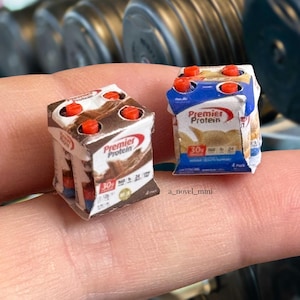 May include: Two miniature boxes of Premier Protein powder, one chocolate and one vanilla, with red lids. The boxes are sitting on a hand.