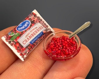 May include: A small bowl of red cranberries in a clear glass bowl with a silver spoon. The bowl is sitting on a hand. A package of Ocean Spray Fresh Premium Cranberries is in the background.