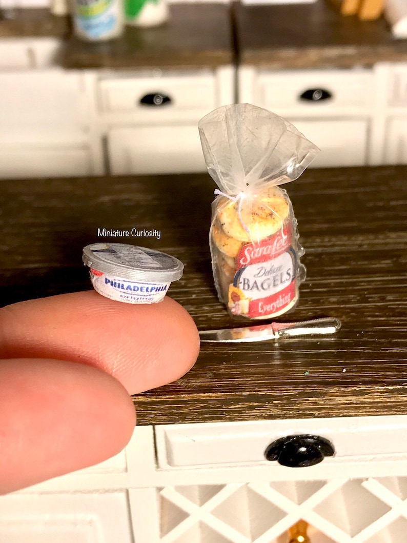 May include: A miniature plastic container of Philadelphia cream cheese next to a bag of mini everything bagels. The bag is tied with a string and has a red and white label that says "Strafel's Everything Bagels".