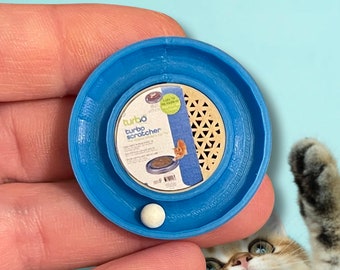 May include: A blue cat scratching post with a white ball in the center. The scratching post has a cardboard insert with a honeycomb pattern and the words "turbo scratcher" printed on it. A cat is looking at the scratching post.