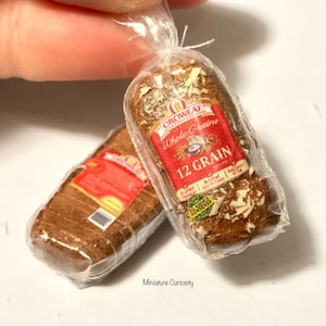 Puede incluir: Dos hogazas de pan en miniatura en bolsas de plástico. Las hogazas son marrones y tienen una cobertura blanca y escamosa. La bolsa de la derecha tiene una etiqueta roja que dice "Oroweat Whole Grains 12 Grain".