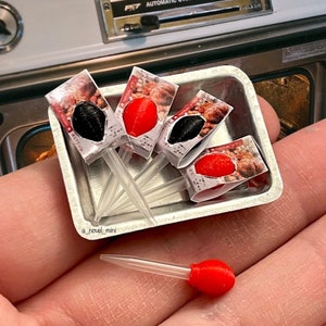 Puede incluir: Un juego de cinco moldes de silicona en miniatura de color rojo y negro con forma de comida en palos de plástico transparente. Los moldes están en una bandeja plateada. La bandeja está en una mano.
