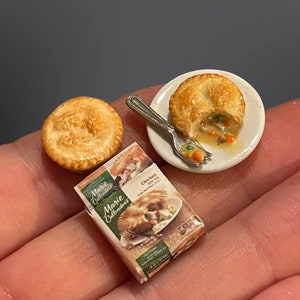 May include: A miniature chicken pot pie on a white plate with a fork. The pie is partially eaten and the filling is visible. There is a whole miniature chicken pot pie next to the plate and a box of Marie Callender's Chicken Pot Pie.