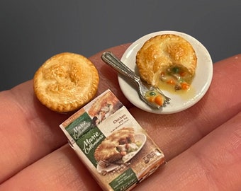 May include: A miniature chicken pot pie on a white plate with a fork. The pie is partially eaten and the filling is visible. There is a whole miniature chicken pot pie next to the plate and a box of Marie Callender's Chicken Pot Pie.