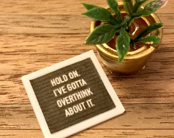 May include: White framed sign with the text "Hold on. I've gotta overthink about it." on a dark brown background. A small potted plant with green leaves is in the background.