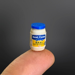 Może przedstawiać: Miniaturowy plastikowy słoiczek z majonezem Best Foods Real. Słoiczek jest biały z niebieską pokrywką i żółtą etykietą z nazwą marki i nazwą produktu.