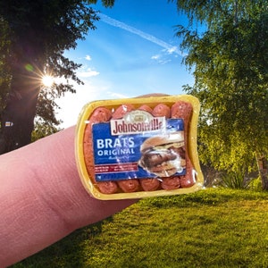 Könnte beinhalten: Eine Miniatur-Plastikverpackung von Johnsonville Brats Original Wurst. Die Verpackung ist gelb mit einem blauen Etikett und einem Bild einer Bratwurst im Brötchen.