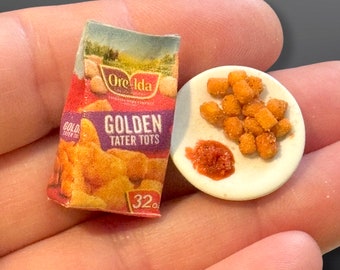 May include: A miniature replica of a box of Ore-Ida Golden Tater Tots, with a small plate of tater tots and a red sauce. The box is red, white, and purple with the words "Ore-Ida" and "Golden Tater Tots" printed on it. The plate is white with a small amount of red sauce in the center.