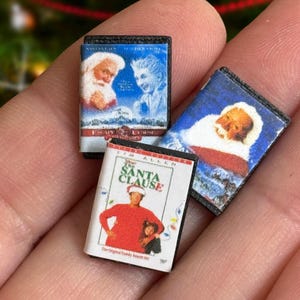 Peut inclure: Miniatures de boîtiers de films sur le thème des fêtes. Les boîtiers ont des dos noirs et des couvertures colorées avec des images du Père Noël et d'autres personnages. Un boîtier indique "The Santa Clause".