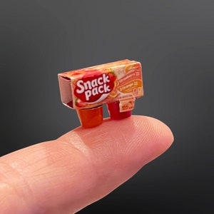 May include: A miniature plastic box of Snack Pack strawberry and orange flavoured pudding cups. The box is red, white and blue with the words "Snack Pack" printed on the front.