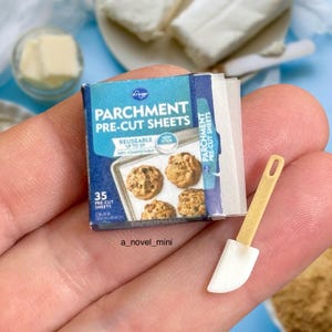 May include: A miniature box of parchment paper baking sheets with a small spatula. The box is blue and white with the text "Parchment Pre-Cut Sheets" and "Reusable Up to 3x". The spatula is white with a brown wooden handle.