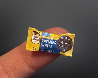 May include: Miniature package of Nestle Toll House Premier White Morsels, 12 oz. The yellow and blue package features a chocolate chip cookie with white chocolate chips. The package is held by a finger.