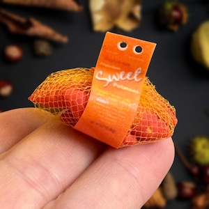 Puede incluir: Una pequeña bolsa de malla llena de batatas rojas, sostenida en una mano. La bolsa está asegurada con una etiqueta naranja que dice "Sweet Potatoes" en escritura blanca. El fondo está borroso, con indicios de follaje otoñal.