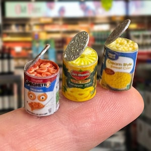 Può includere: Tre lattine di cibo in miniatura, tra cui pezzi di ananas Del Monte, SpaghettiOs e una lattina di mais. Le lattine sono su un dito, mostrando la loro piccola dimensione.