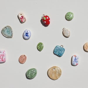 May include: Assortment of ceramic charms with various designs, including leaves, flowers, a ladybug, and a swirl pattern. The charms are in shades of green, blue, red, and white.
