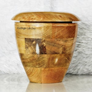 May include: A wooden urn with a lid, made of multiple pieces of wood with a natural wood finish. The urn is in the shape of a vase and has a label that reads "Vintage Life Inc."