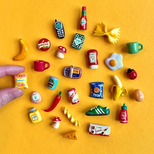 Pode incluir: Uma coleção de alimentos e objetos em miniatura, incluindo uma banana, uma pimenta vermelha, um tênis verde, um telemóvel, uma garrafa de molho picante, um saco de batatas fritas e uma barra de chocolate Kinder Bueno, sobre um fundo amarelo.