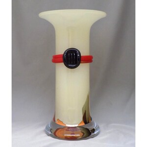 Könnte beinhalten: Eine hohe, cremefarbene Glasvase mit ausgestelltem Oberteil und klarem, abgerundetem Boden. Ein rotes Band mit einem schwarzen, oval geformten Akzent umschließt die Vase. Die Vase ist etwa 30 cm hoch.