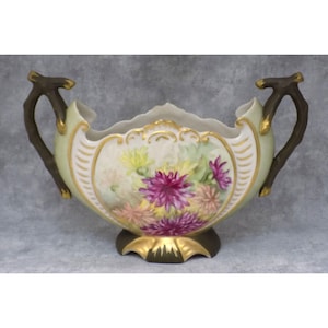 May include: A decorative porcelain bowl with a floral design. The bowl has a light green background, gold trim, and two brown handles. The floral design includes pink, purple, and yellow flowers. The bowl sits on a dark green base.