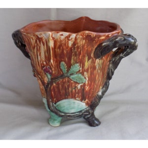 May include: A brown ceramic planter with three legs and two handles shaped like tree branches. The planter is decorated with a green and red vine with leaves and flowers.