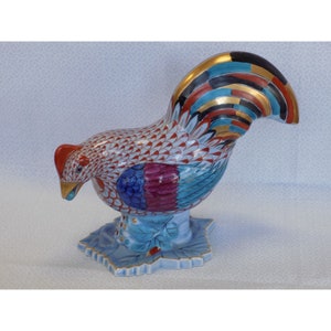 May include: A colorful porcelain rooster figurine with a blue, red, and gold painted design. The rooster is standing on a blue and white leaf-shaped base.
