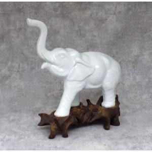 May include: A white ceramic elephant ornament with its trunk raised, set on a dark brown, textured wooden base. The elephant has detailed features and a glossy finish. The ornament is a decorative item.
