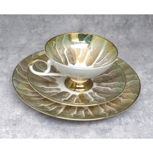 Puede incluir: Un juego de té vintage que incluye una taza, un platillo y un plato de postre. El juego tiene una base blanca con un diseño geométrico dorado y verde. La taza y la base tienen detalles dorados. El juego es para una persona.