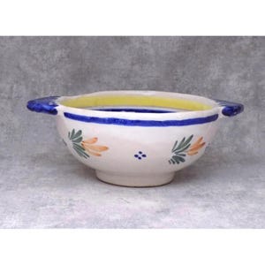 May include: A white ceramic bowl with blue and yellow accents. The bowl features a blue stripe and handles, with floral designs in orange and green. The interior rim is yellow.