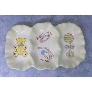 Puede incluir: Un plato de cerámica blanco para bebé con tres secciones. La sección central tiene el texto "Babies are the sweetest things" con un imperdible rosa y azul. La sección izquierda tiene un osito de peluche amarillo con una pajarita azul. La sección derecha tiene un sonajero verde y amarillo.