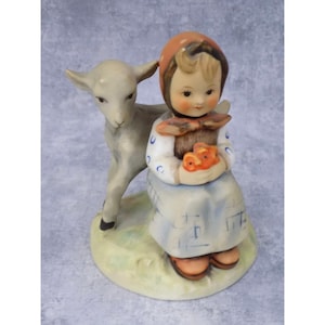 May include: A ceramic figurine depicting a young girl in a bonnet and dress, holding a basket of flowers, with a lamb standing beside her. The figurine is painted in soft colors and has a matte finish.