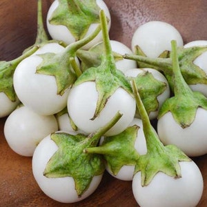 May include: A bowl of white eggplants with green stems. The eggplants are arranged in a cluster, with some of them overlapping.