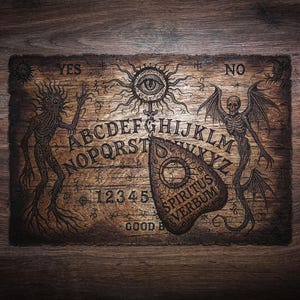 May include: A rectangular wooden spirit board featuring an engraved alphabet, numbers 1-5, and the words YES, NO, and GOOD BYE. The board displays illustrations of a sun with an eye, a skeletal winged creature, and a spindly figure, with a planchette labelled SPIRITUS VERBUM.