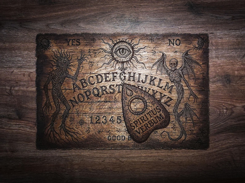May include: A rectangular wooden spirit board featuring an engraved alphabet, numbers 1-5, and the words YES, NO, and GOOD BYE. The board displays illustrations of a sun with an eye, a skeletal winged creature, and a spindly figure, with a planchette labelled SPIRITUS VERBUM.