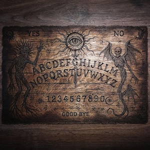 May include: A rectangular wooden spirit board featuring an engraved alphabet from A to Z, numbers 0 through 9, and the words YES, NO, and GOOD BYE. The board displays intricate illustrations of a central eye, a skeletal figure, and a creature.