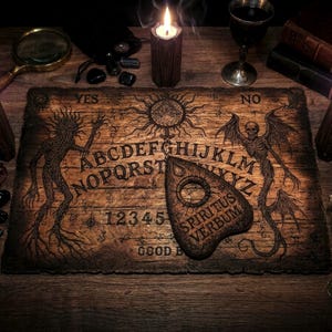 May include: A rectangular wooden spirit board featuring an engraved alphabet, numbers 1-9 and 0, the words YES, NO, and GOOD BYE, and illustrations of a sun and two winged figures. A matching wooden planchette with the text SPIRITUS VERBUM rests on top.