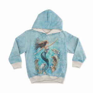 May include: A light blue hoodie featuring a mermaid design. The illustration shows a mermaid with flowing brown hair and a teal tail, surrounded by seals. The hoodie has a light blue hood and cuffs, with a cream-colored lining.
