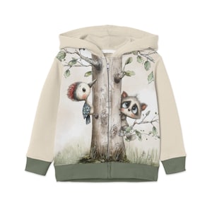 Könnte beinhalten: Beige Kapuzenjacke mit einer skurrilen Illustration eines Spechts und eines Waschbären, die hinter einem Baum hervorblicken. Die Ärmel und der Besatz sind olivgrün. Das Design hat einen Aquarellstil.